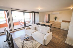 Un salon moderne avec un canapé, une table basse et un téléviseur. Il y a un balcon avec une chaise et une table. On aperçoit la cuisine au fond, avec une table à manger et des chaises.