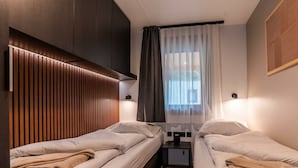 A compact hotel room with two single beds, a small bedside table, a wall-mounted light, and a window with sheer curtains. - Wulfenia Waterfront 6 (Ossiach)