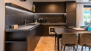 A modern kitchen with dark cabinetry, a marble countertop, and a dining area with a wooden table and chairs. - Wulfenia Waterfront 6 (Ossiach)