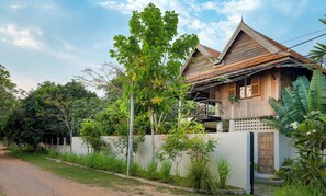 A traditional wooden house with a balcony, surrounded by lush greenery and a clear sky.