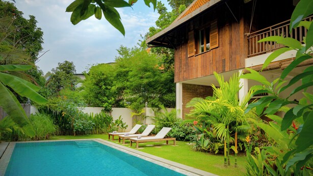 A pool with lounge chairs, surrounded by lush greenery and a wooden building. - Beautiful Khmer house with pool at the edge of the Angkor conservation zone (ក្រុងសៀមរាប)