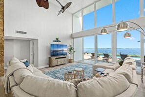 Ein geräumiges Wohnzimmer mit großem Fernseher, bequemen Sitzmöglichkeiten und Meerblick durch große Fenster.