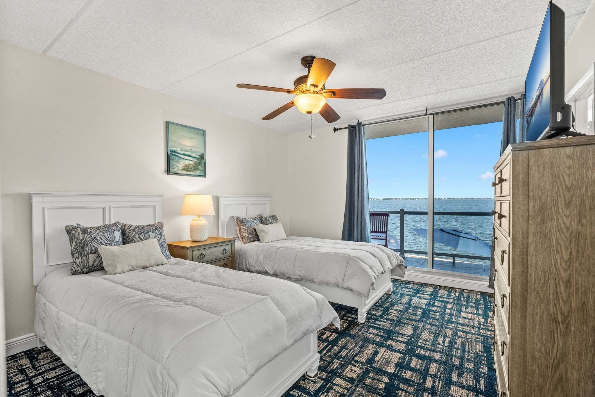 Ein Hotelzimmer mit zwei Betten, einem Deckenventilator, einem großen Fenster mit Blick auf das Meer und einer Holzkombination.
