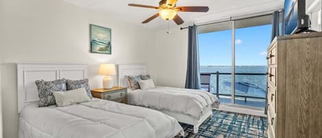 Ein Hotelzimmer mit zwei Betten, einem Deckenventilator, einem großen Fenster mit Blick auf das Meer und einer Holzkombination.