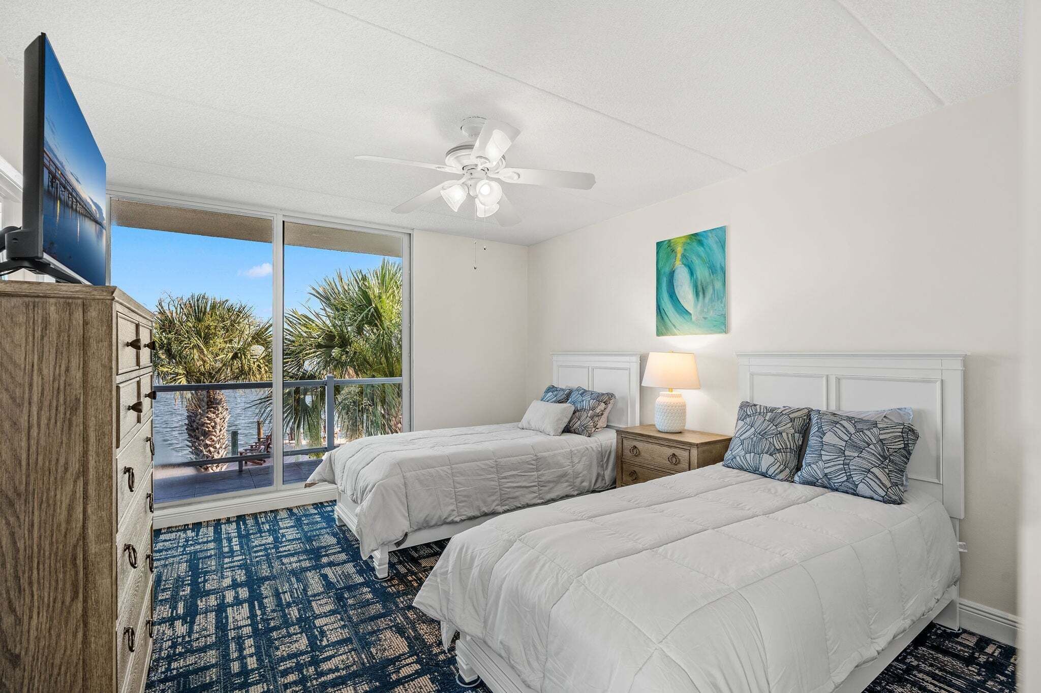 A bedroom with two beds, a ceiling fan, a TV, a balcony with palm trees, and a painting on the wall.
