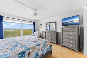 Un dormitorio con cama, cómoda, silla, televisor y un amplio ventanal con vistas.