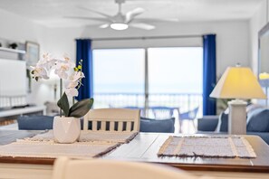 A modern living room with a dining area, a ceiling fan, a view through large windows, and a lamp on a side table. - Stunning Oceanfront Condo W/ Private Balcony & Heated Pool! 🌊 (Cocoa Beach)
