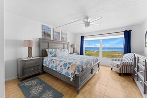 Ein Schlafzimmer mit einem Bett, einem Nachttisch, einer Kommode, einem Sessel sowie einem Deckenventilator und einem großen Fenster mit Aussicht.