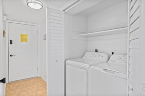 Un cuarto de lavandería con una puerta blanca, un estante en la pared y dos lavadoras de carga frontal apiladas.