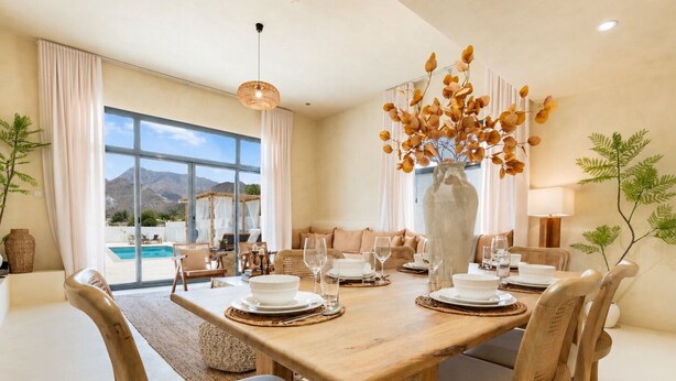A dining area with a large table set for four, a vase with dried flowers, and a view of a pool and mountains.
