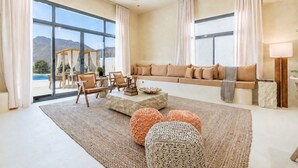 A spacious living room with a large sofa, a coffee table, and a view of a swimming pool and mountains.