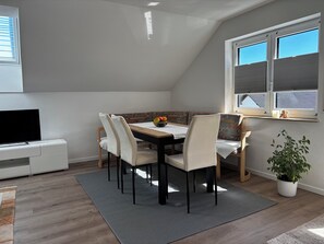 Een eetgedeelte met een tafel, stoelen, een televisie, een potplant en een raam met zonwering.