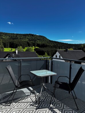 Ein Balkon mit Glastisch und zwei Stühlen, mit Blick auf einen bewaldeten Hügel.