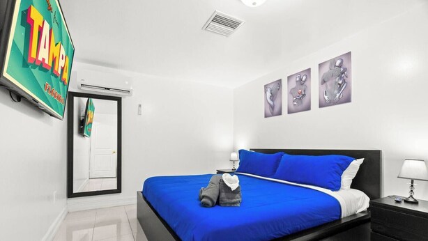 A hotel room with a bed, a nightstand, a wall-mounted TV, and artwork. - Cozy Apt, King Bed + Huge TV, Near TPA & Stadium (Tampa)