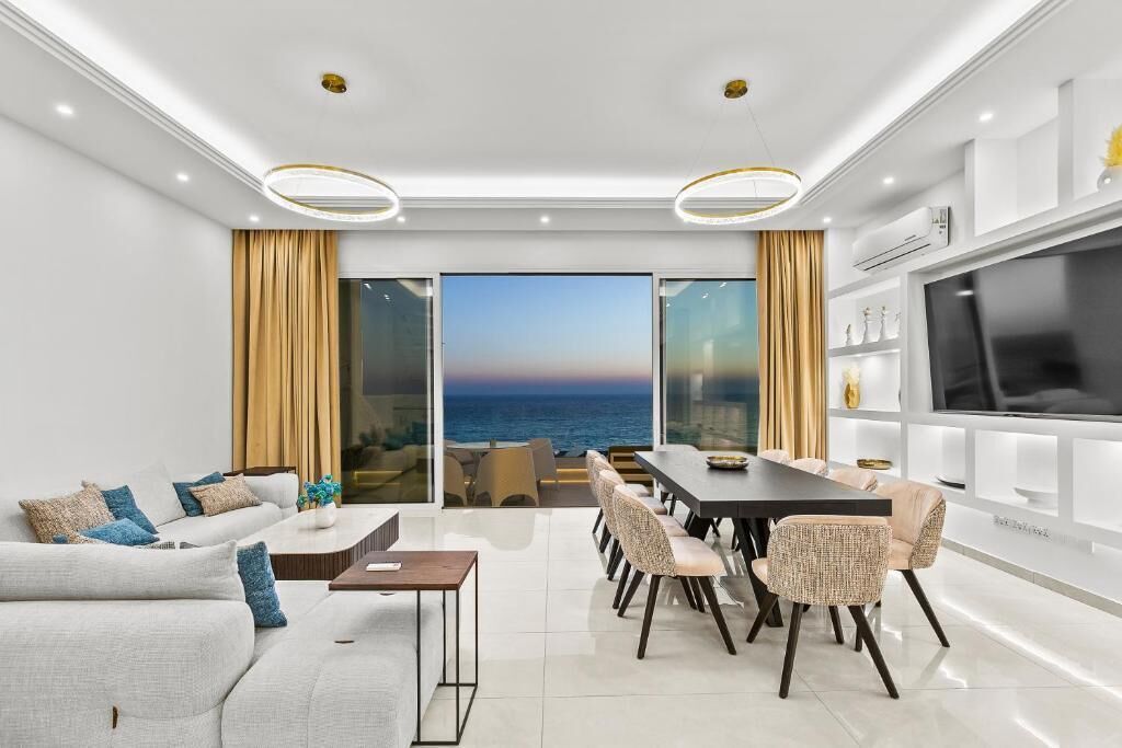 A modern living room with a large TV, a dining area, and a view of the sea.