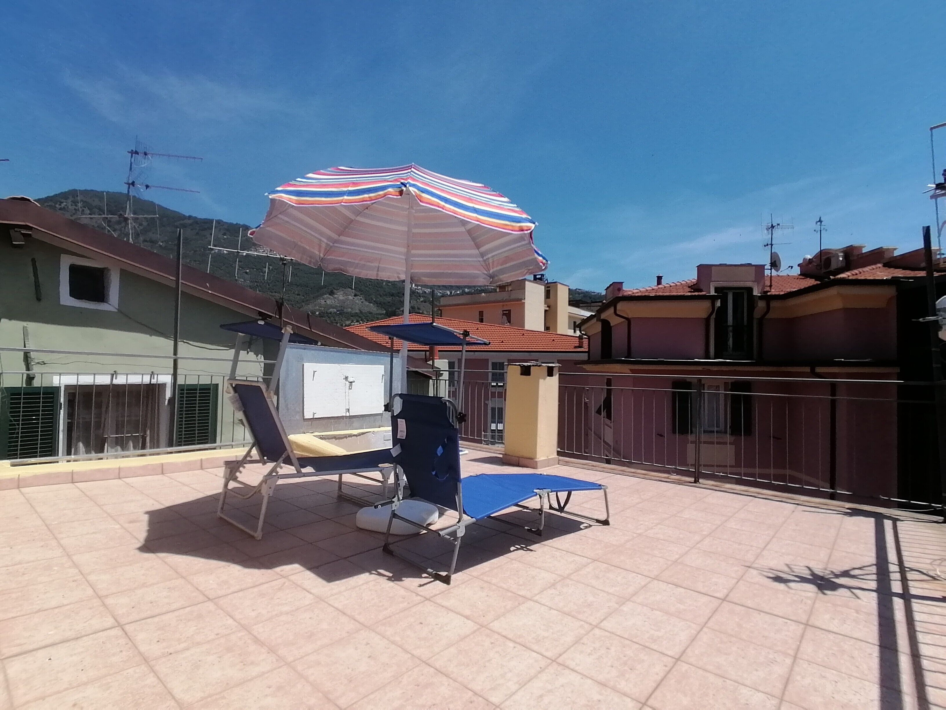 Une terrasse sur le toit avec un parasol à rayures, deux chaises longues, un tapis bleu et un tableau blanc.