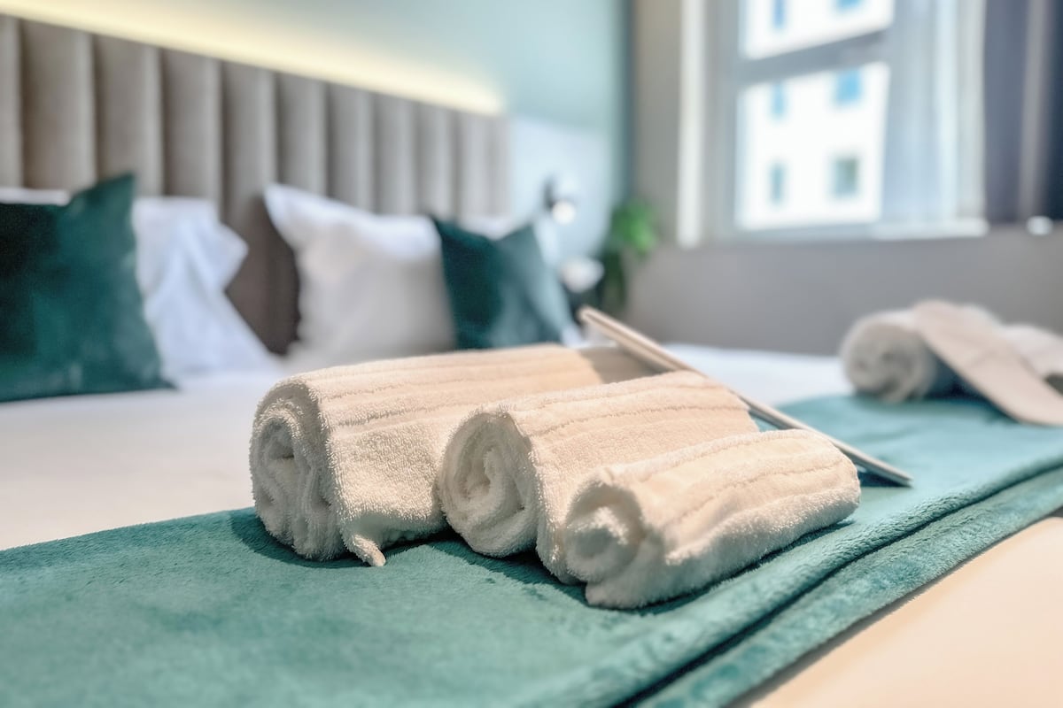 A neatly made bed with rolled white towels, a teal throw blanket, and a wooden bed frame in a hotel room.
