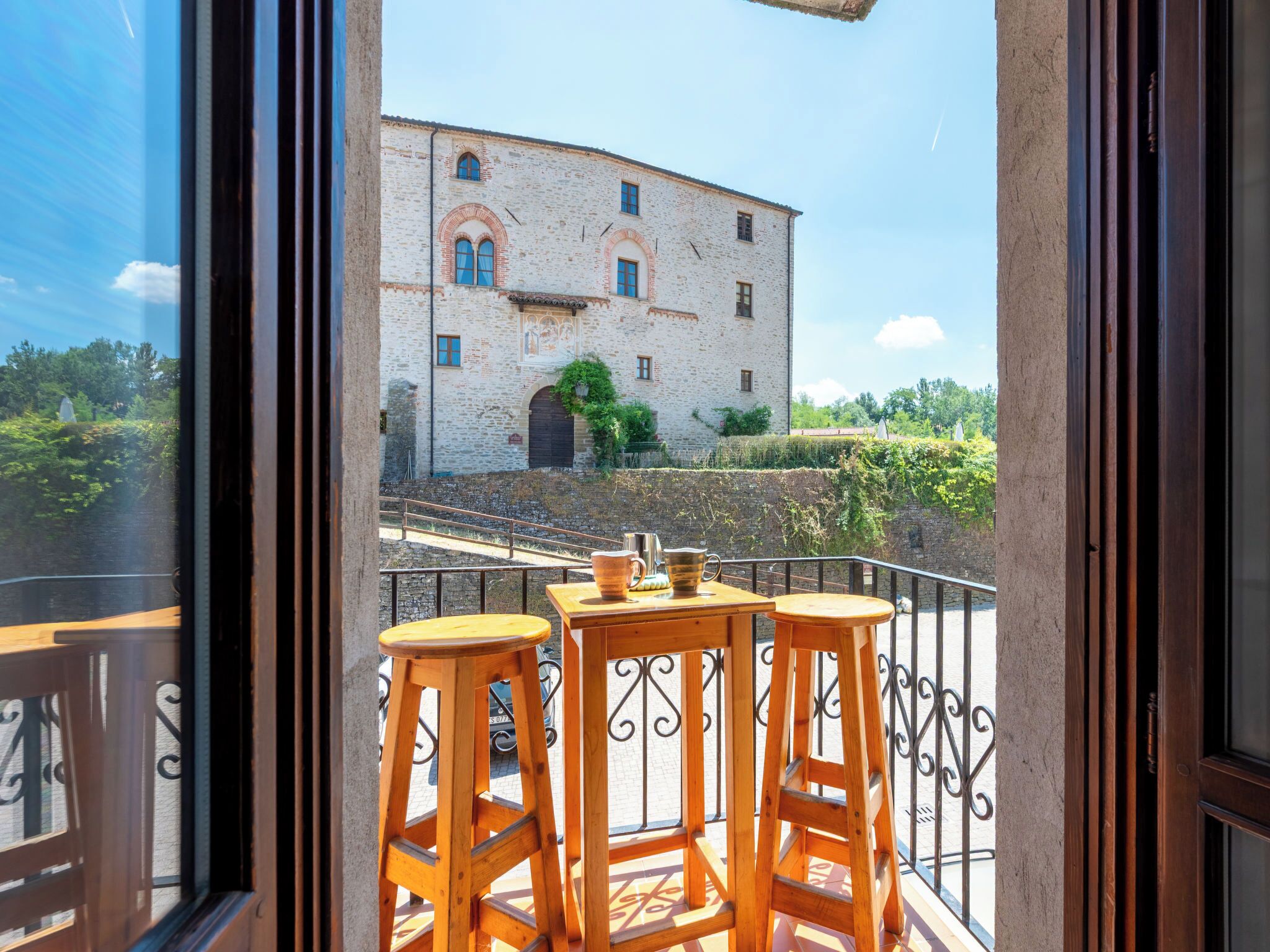 A balcony with wooden stools and a table, overlooking a historic building with arched windows and a stone wall.