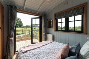 A bedroom with a large bed, a window with a view of a fenced outdoor area, and a radiator. - Luxury Shepherds Hut with hot tub, firepit and gas barbeque (Anglesey)