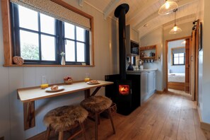 A cozy kitchen with a wood-burning stove, a dining table with chairs, and a window with a view of trees. - Luxury Shepherds Hut with hot tub, firepit and gas barbeque (Anglesey)
