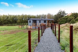 A pathway leading to a small, single-story building with a corrugated metal roof, surrounded by a wire fence and gravel. - Luxury Shepherds Hut with hot tub, firepit and gas barbeque (Anglesey)