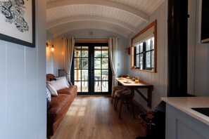 A cozy room with a wooden floor, a brown sofa, a dining table with chairs, and a large window with curtains. - Luxury Shepherds Hut with hot tub, firepit and gas barbeque (Anglesey)