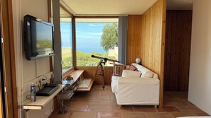 Ein Zimmer mit Fernseher, Teleskop, weißem Sofa, Holzwandverkleidung und Blick ins Freie.