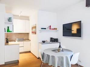 Eine kleine Küche mit weißen Schränken, einem Esstisch mit Tischdecke und einem an der Wand befestigten Flachbildfernseher.