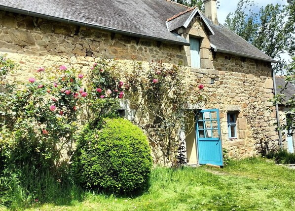 Un edificio in pietra con una porta blu, una piccola finestra e un giardino con fiori rosa.