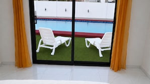 Quarto com uma cama, duas cadeiras brancas, uma porta de vidro deslizante e vista para uma piscina.