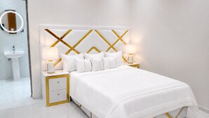 Um quarto moderno com uma cama branca, detalhes dourados, uma mesa de cabeceira com luminária e um banheiro com espelho e pia.
