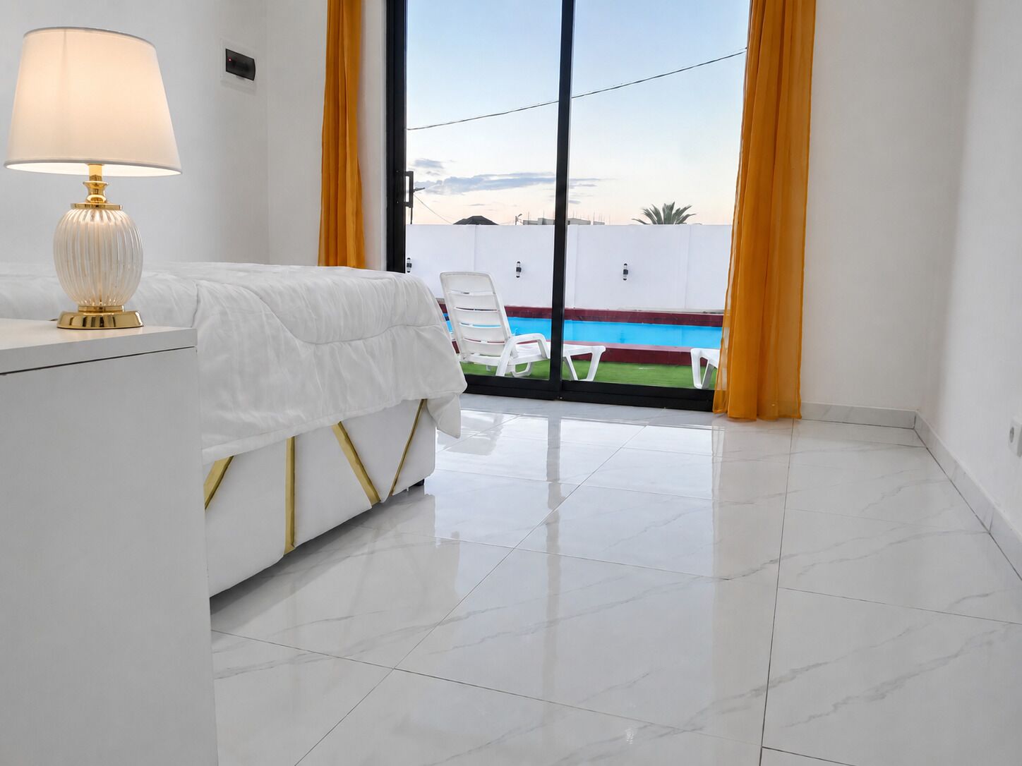 Habitación de hotel con una cama, una mesita de noche con lámpara, vista a una piscina a través de un ventanal de vidrio y piso de baldosas.