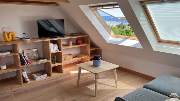 Ein Loft-Zimmer mit schrägem Dach, Holzböden, einem Fernseher, einem kleinen Tisch und Blick auf eine Stadtlandschaft durch ein Oberlicht.