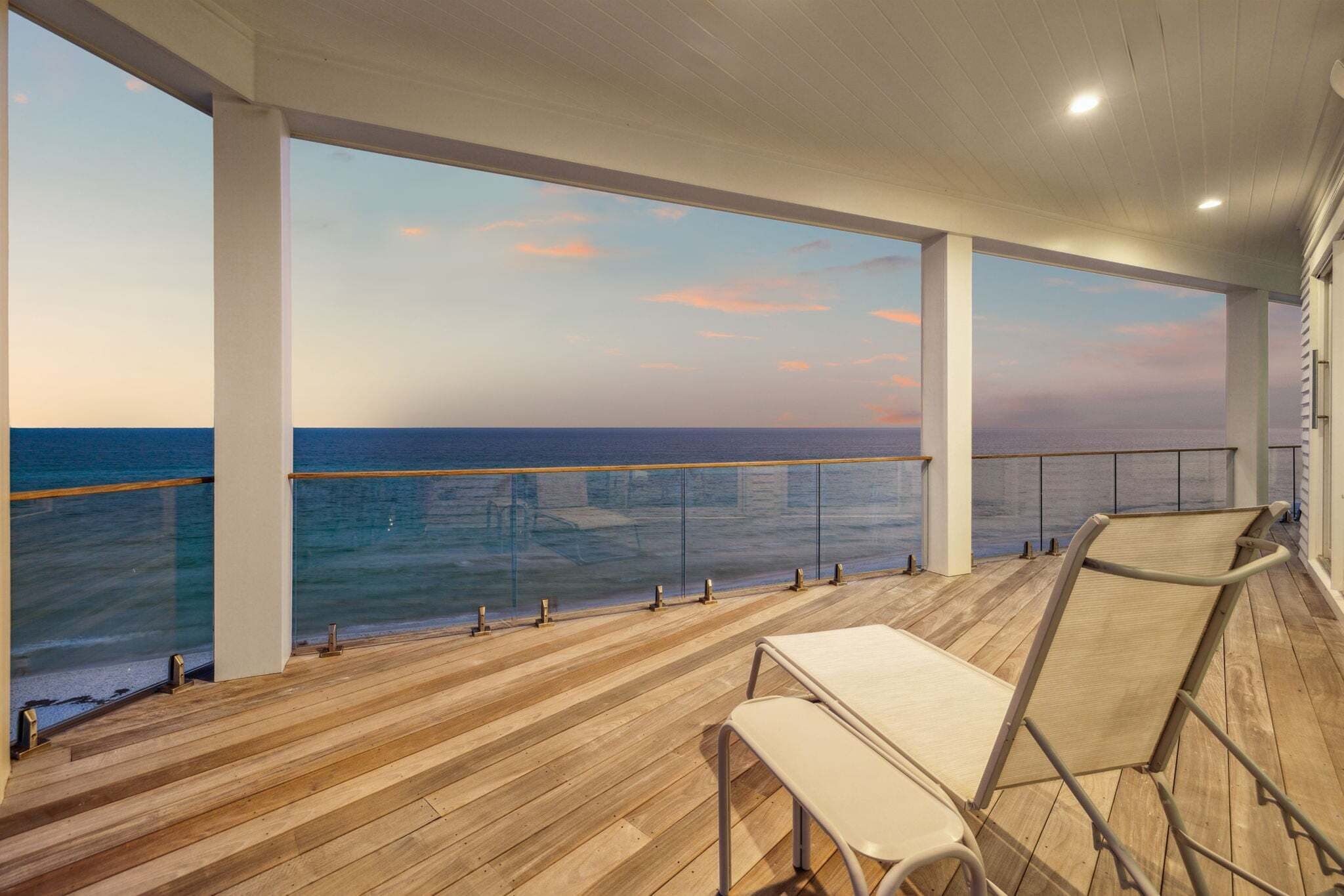 Uma área externa coberta com deck de madeira, guarda-corpo de vidro e vista para o mar.