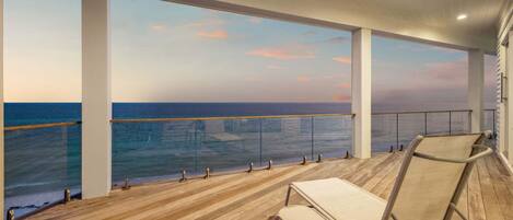 Uma área externa coberta com deck de madeira, guarda-corpo de vidro e vista para o mar.