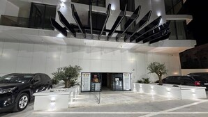 Entrada de um edifício moderno com área de estacionamento, dois carros estacionados e uma árvore ao primeiro plano.