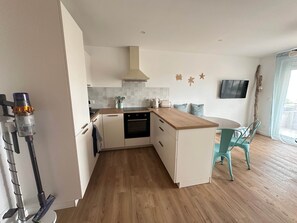 A modern kitchen with white cabinets, a wooden countertop, and a built-in oven. There is a dining area with a round table and teal chairs. - Fully renovated apartment with a beautiful ocean view (Sausset-les-Pins)
