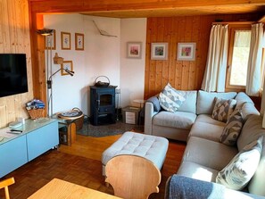 A cozy living room with a sofa, ottoman, and a television. There is a fireplace and a small table with a lamp.