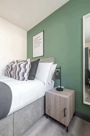 Un dormitorio con cama, almohadas, mesita de noche, lámpara, una cita enmarcada en la pared y espejo.