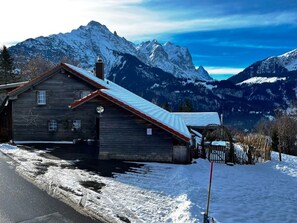 A wooden cabin with a sloped roof, a chimney, and a window, set against a backdrop of snow-covered mountains and clear skies. - HasliRent, Lindeli, (Hasliberg Reuti), by Interhome (Hasliberg)