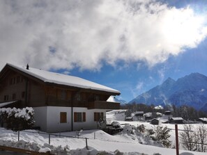 Eine zweistöckige Chalet mit schneebedecktem Dach, vor einer Berglandschaft und einem teils bewölkten Himmel gelegen.