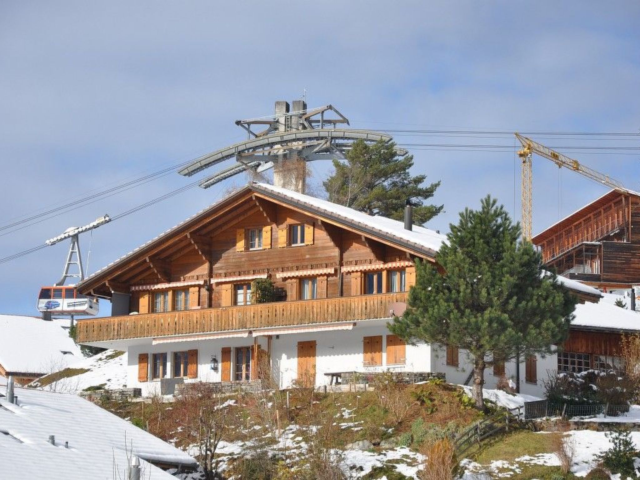 Eine Holzhütte mit Balkon, im Hintergrund eine Seilbahn und eine verschneite Landschaft.