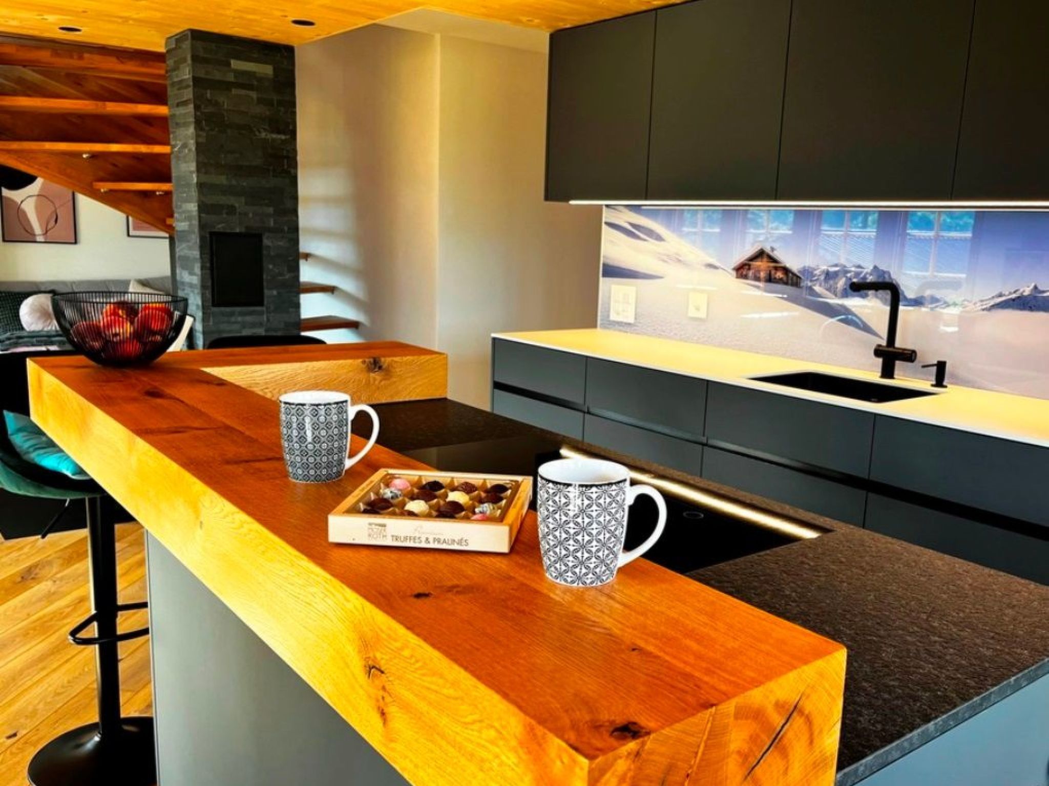 A modern kitchen with a wooden bar counter, two patterned mugs, a box of chocolates, and a view of a snowy landscape through a window.