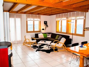 Una sala de estar con techo de madera, un sofá negro con cojines, dos mecedoras, una mesa de centro y una mesa de comedor con sillas.