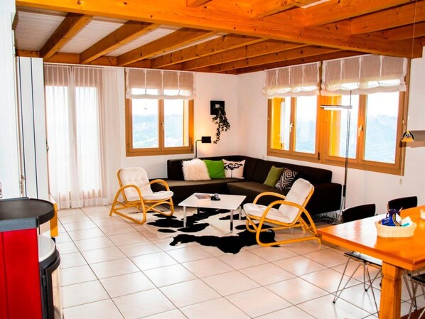 A living room with a wooden ceiling, a black sofa with cushions, two rocking chairs, a coffee table, and a dining table with chairs.