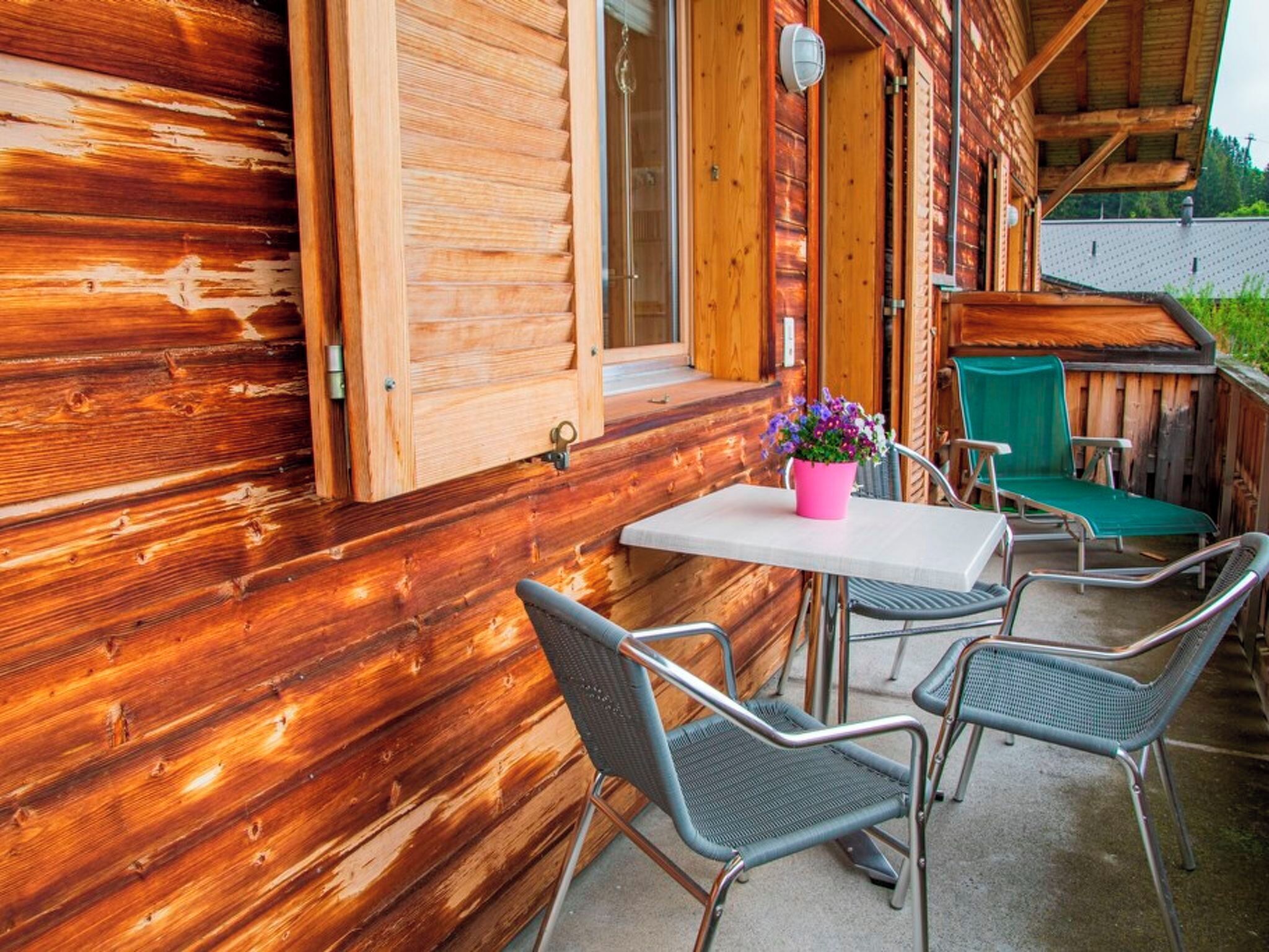 Une cabane en bois dotée d’un patio comprenant une table et des chaises, une plante en pot et un fauteuil vert.