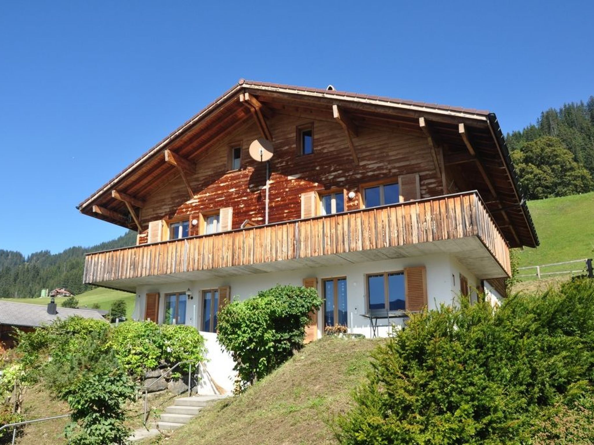 A two-story wooden chalet with a balcony, surrounded by greenery and a clear sky.