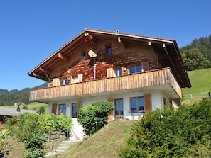 Un chalet de madera de dos pisos con un balcón, rodeado de vegetación y un cielo despejado.