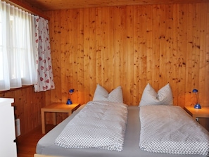 Ein Zimmer mit Holzwandverkleidung, zwei Betten mit gemusterten Bettbezügen, einem Nachttisch mit Lampe und einem Fenster mit durchsichtigen Vorhängen.