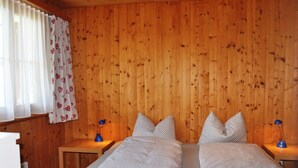 Ein Zimmer mit Holzwandverkleidung, zwei Betten mit gemusterten Bettbezügen, Nachttischlampen und einem Fenster mit durchsichtigen Vorhängen.